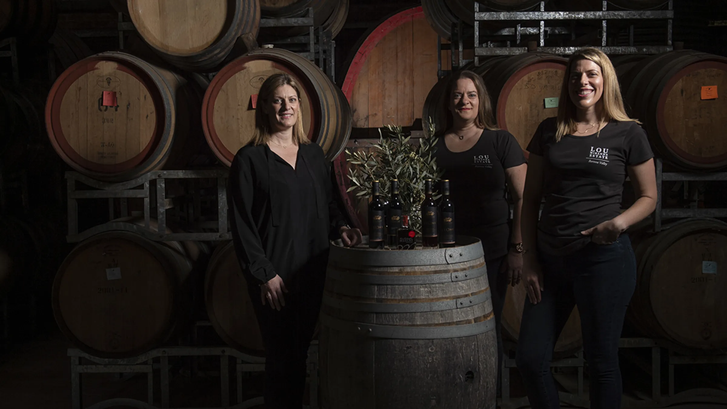 Three persons in front of wood barrels with a barrel as a table and bottles of wine